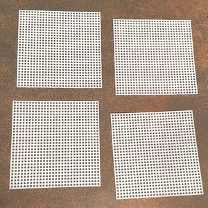 May include: Four white plastic canvas squares with a grid pattern. The squares are arranged on a brown surface. These craft supplies are used for needlepoint, cross-stitch, and other DIY projects.