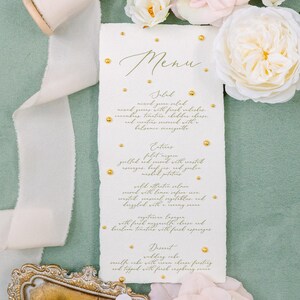 May include: A wedding menu with gold lettering and accents, surrounded by white and pink roses. The menu lists salad, entrees, and dessert options. A spool of ribbon and a decorative tray are also present.
