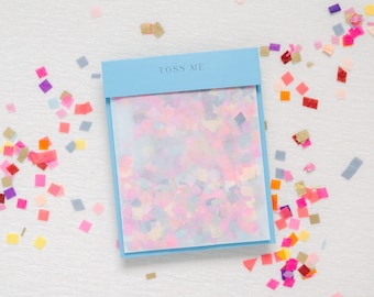May include: A blue box with the text "Toss Me" on the front. The box contains a clear plastic bag filled with colorful confetti.