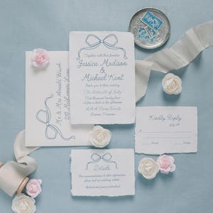 May include: Wedding invitation suite with a blue and white color scheme. The invitation features a hand-drawn bow and the text "Together with their families, Jessica Madison & Michael Kent invite you to their wedding. The thirteenth of July, Two thousand twenty-four at four o'clock in the afternoon. A A A Farm Events,  Lanna, Tennessee". The RSVP card has the text "Kindly Reply by July 1st". The details card has the text "For accommodations & other information please visit our wedding website theknot.com/jessicamikhael".