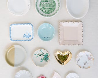 May include: A collection of fourteen small ceramic and porcelain dishes in various shapes and colors. The set includes round, square, heart-shaped, and petal-shaped plates featuring floral patterns, solid colors, gold accents, and a green building print.