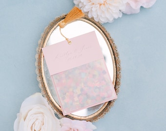 May include: A pink envelope with a white tassel, filled with confetti, sits on a gold oval tray with a mirror bottom. The tray is surrounded by white and pink flowers.