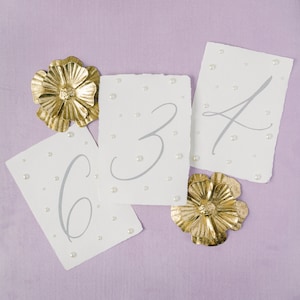May include: Three white cards with the numbers 6, 3, and 4 written in gray on them. The cards are decorated with small white pearl-like embellishments. Two gold paper flowers are on the cards.
