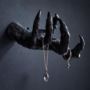 May include: A black wall-mounted claw-shaped jewelry holder with textured skin details. A thin gold-toned chain necklace with an oval blue pendant and a small hoop earring with a star-shaped charm are hanging from the fingers.