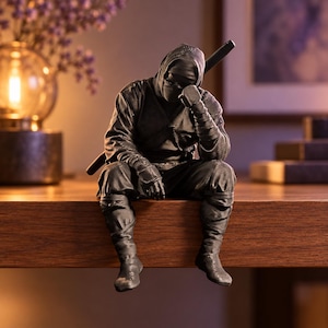 May include: A matte black ninja figurine in a seated position with legs dangling. The figure wears a traditional hooded mask and wraps, with a katana sword strapped across the back. One hand rests on the knee while the other touches the forehead.