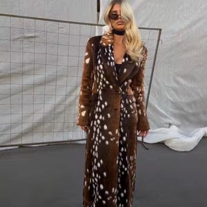 May include: A long, calf-length coat featuring a brown and white spotted pattern reminiscent of a fawn's coat. The garment has a notched lapel collar, long sleeves, and a button-front closure. The texture appears soft and plush throughout.