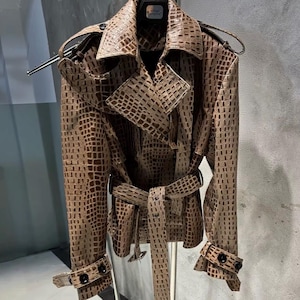 Puede incluir: Una gabardina de color tostado con un patrón texturizado de piel de cocodrilo en toda la prenda. El abrigo incluye un cuello ancho, charreteras en los hombros, mangas largas con puños de hebilla y un cinturón a juego con hebilla y relieve.