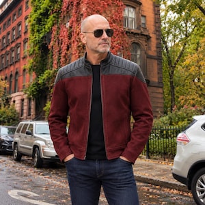 Puede incluir: Una chaqueta de ante color granate con paneles de cuero negro en los hombros, cuello alzado y cremallera frontal. La prenda se lleva sobre una camiseta negra lisa y pantalones vaqueros azul oscuro, junto con gafas de sol tipo aviador.