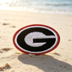 May include: An oval-shaped embroidered patch featuring a black background with a bold white letter G in the center, surrounded by a thick red border. The design is stitched with high-contrast thread, creating a textured appearance on the fabric.