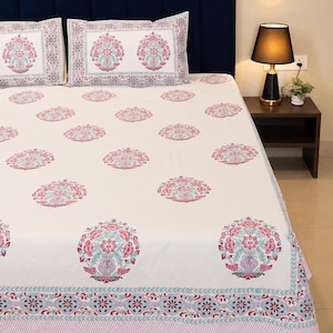 Op de afbeelding: Een wit laken en twee bijpassende kussenslopen met een herhalend bloemenpatroon in roze, turquoise en grijstinten. De stof heeft een decoratieve rand met een geometrisch ontwerp in roze en turquoise aan de onderkant.