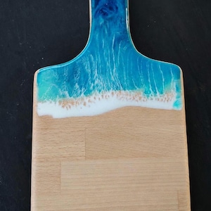 May include: A rectangular wooden serving board featuring a handle with a hole at the top. The upper portion is coated in a glossy resin finish with blue and turquoise swirls and white foam detailing, resembling ocean waves hitting a sandy beach.