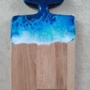 May include: A rectangular wooden serving board featuring a handle shaped like a whale tail. The top portion of the board is coated in a glossy resin finish with swirling shades of deep blue, turquoise, and white, mimicking ocean waves.