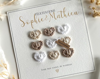 Peut inclure: Une carte carrée blanche avec le texte doré "Félicitations Sophie & Mathieu" et "Tous nos voeux de bonheur" avec un petit cœur. Neuf petits cœurs crochetés en blanc, beige et marron sont fixés, dont un avec une étiquette marron "Douces pensées".