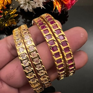 May include: Four gold-toned bangles featuring a channel-set design. Two bangles are inlaid with square-cut clear stones, while the other two bangles are inlaid with square-cut magenta stones. The metal bands have a polished finish.