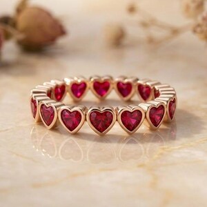 Puede incluir: Un anillo de eternidad en oro rosa con una banda continua de piedras preciosas rojas en forma de corazón. El anillo está diseñado con una serie de engastes en forma de corazón conectados, cada uno con una piedra roja facetada.