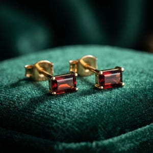 May include: A pair of stud earrings featuring rectangular baguette-cut deep red garnet gemstones set in polished yellow gold metal. Each earring has a secure post back and a clean, geometric design that highlights the vibrant red colour of the stones.