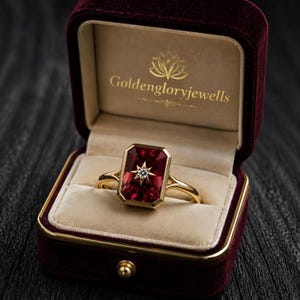 May include: A gold ring featuring a rectangular red gemstone with a central star-shaped setting containing a small clear stone. The ring is displayed inside a dark red velvet box with the gold text "goldengloryjewells" printed on the inner lid.