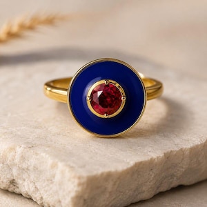 Op de afbeelding: Een gouden ring met een cirkelvormig middendeel met een diepblauwe emaille inleg rondom een centrale ronde gefacetteerde rode edelsteen, vastgehouden door vier kleine pootjes. De ringband is van glad en gepolijst goud, wat een opvallend contrast creëert tussen de levendige rode steen, het donkerblauwe emaille en de metalen gouden zetting.