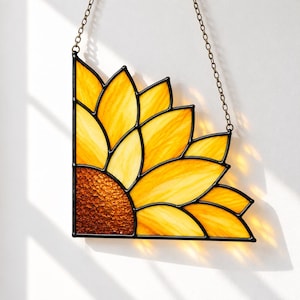 Könnte beinhalten: Ein Sonnenblumen-Eckelement aus Buntglas mit leuchtend gelben Blütenblättern und einer strukturierten bernsteinfarbenen Mitte. Das Design ist in schwarzes Metall gefasst und verfügt über eine dünne Metallkette zum Aufhängen als Sonnenfänger.