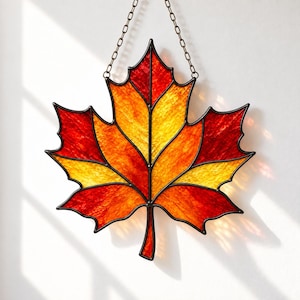 Könnte beinhalten: Ein Ahornblatt-Sonnenfänger aus Buntglas mit strukturierten Glassegmenten in Rot-, Orange- und Gelbtönen. Das Blatt ist mit dunklen Metalllötlinien umrandet und an einer dünnen Metallkette befestigt, die an den oberen Spitzen angebracht ist.