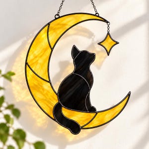 Könnte beinhalten: Ein Buntglas-Sonnenfänger mit einem gelben Halbmond, auf dem eine schwarze Katzensilhouette sitzt. Ein kleiner, vierzackiger gelber Stern hängt an einer Metallkette an der oberen Spitze des Mondes. Das Objekt ist an einer Kette aufgehängt.