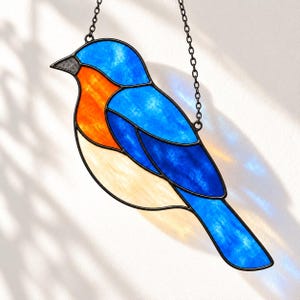 Könnte beinhalten: Ein Buntglas-Sonnenfänger in Form eines Blauvogels mit leuchtend blauen, hellorangen und cremefarbenen Glassegmenten, die durch dunkle Metalllötlinien verbunden sind und an einer dünnen schwarzen Metallkette hängen.