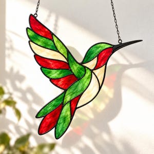Könnte beinhalten: Ein Kolibri-Sonnenfänger aus Buntglas mit leuchtend roten, grünen und cremefarbenen Glassegmenten, die mit schwarzem Metall umrandet sind. Der Vogel ist im Flug mit ausgebreiteten Flügeln dargestellt und an einer dünnen Metallkette aufgehängt.