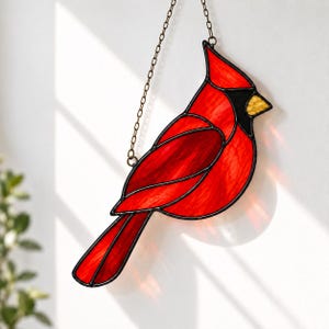 May include: A stained glass suncatcher shaped like a cardinal bird, featuring vibrant red glass panels with black lead lines, a small yellow beak, and a black face mask. The piece hangs from a thin metal chain attached to the top of the bird.