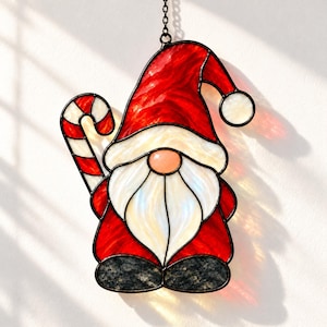 Könnte beinhalten: Ein Buntglas-Suncatcher in Form eines Wichtels mit roter Mütze und weißem Bommel, langem weißem Bart und dunkelgrauen Stiefeln. Der Wichtel hält eine rot-weiß gestreifte Zuckerstange und hängt an einer Metallkette.