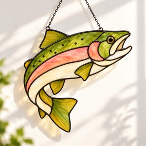 Könnte beinhalten: Ein Sonnenfänger aus Buntglas in Form einer Forelle, bestehend aus grünen, rosafarbenen und cremefarbenen Glassegmenten mit schwarzer Metallumrandung. Der Fisch hängt an einer dünnen schwarzen Metallkette und zeigt einen gebogenen Körper.