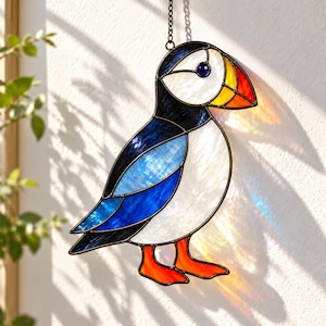 May include: A stained glass suncatcher shaped like a puffin bird. The bird features a white face, a black back, blue wing accents, and a bright orange and yellow beak. It has orange feet and is suspended by a thin metal chain for hanging.