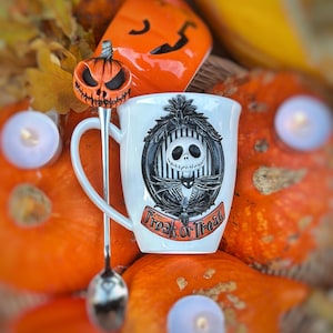 Könnte beinhalten: Ein weißer Keramikbecher mit einem schwarz-weißen Porträt einer skelettartigen Figur in einem Rahmen und dem Text "Trick or Treat" auf einem orangefarbenen Banner, dazu ein silberner Löffel mit einem orangefarbenen Kürbisaufsatz.