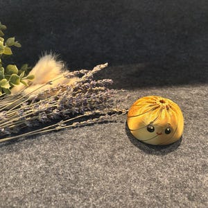Könnte beinhalten: Eine kleine, runde, goldgelbe Knödel-Figur mit gefalteter Oberseite und einem handbemalten Gesicht mit zwei schwarzen Augen und einem kleinen roten Mund, platziert neben einem Bündel getrockneter Lavendelzweige und grüner Eukalyptusblätter.