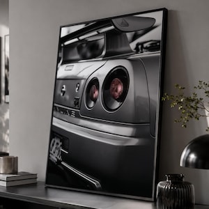 Könnte beinhalten: Ein schwarz-weiß gerahmter Wandkunstdruck, der die Rückansicht eines Sportwagens zeigt. Das Bild hebt die runden Rückleuchten, einen markanten Heckspoiler und das GT-R-Emblem auf dem Kofferraumdeckel in einem kontrastreichen Monochrom-Stil hervor.