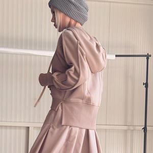 Peut inclure: Une personne porte un sweat à capuche marron clair, un pantalon plissé assorti, un bonnet en tricot gris et des baskets grises. La tenue a une esthétique décontractée mais élégante. Le fond est un mur blanc uni.