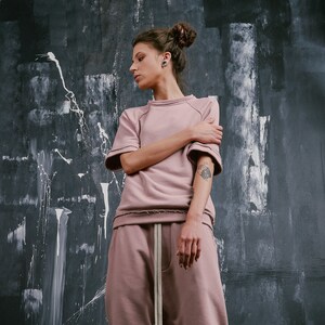 Op de afbeelding: Een vrouw draagt een stoffig roze sweatshirt en een bijpassende joggingbroek met een trekkoord. Ze draagt witte sneakers met oranje en bruine accenten.