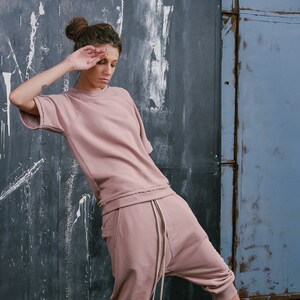 Op de afbeelding: Een vrouw draagt een roze sweatshirt en een bijpassende broek met een trekkoord. Ze draagt sneakers in wit, oranje en blauw.