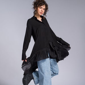 May include: A woman wearing a black long-sleeved shirt with a ruffled hem and light blue jeans. She is wearing black shoes and has curly brown hair.