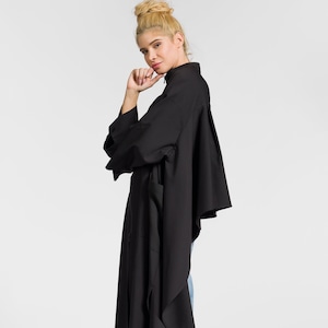 Könnte beinhalten: Ein schwarzes Langarmhemd mit Stehkragen und einem langen, fließenden Saum. Das Hemd ist über den Körper des Models drapiert und erzeugt eine dramatische Silhouette. Das Model trägt eine blaue Jeans und schwarze Stiefel.