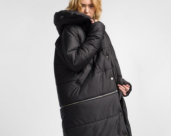 May include: A black long puffer coat with a hood. The coat has a zipper and button closure. The model is wearing the coat with beige pants and black high-top sneakers.