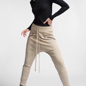 Puede incluir: Una mujer con una camisa negra de manga larga con hombros descubiertos y pantalones beige con cord&oacute;n y bolsillos con cremallera. Lleva zapatillas negras de ca&ntilde;a alta.