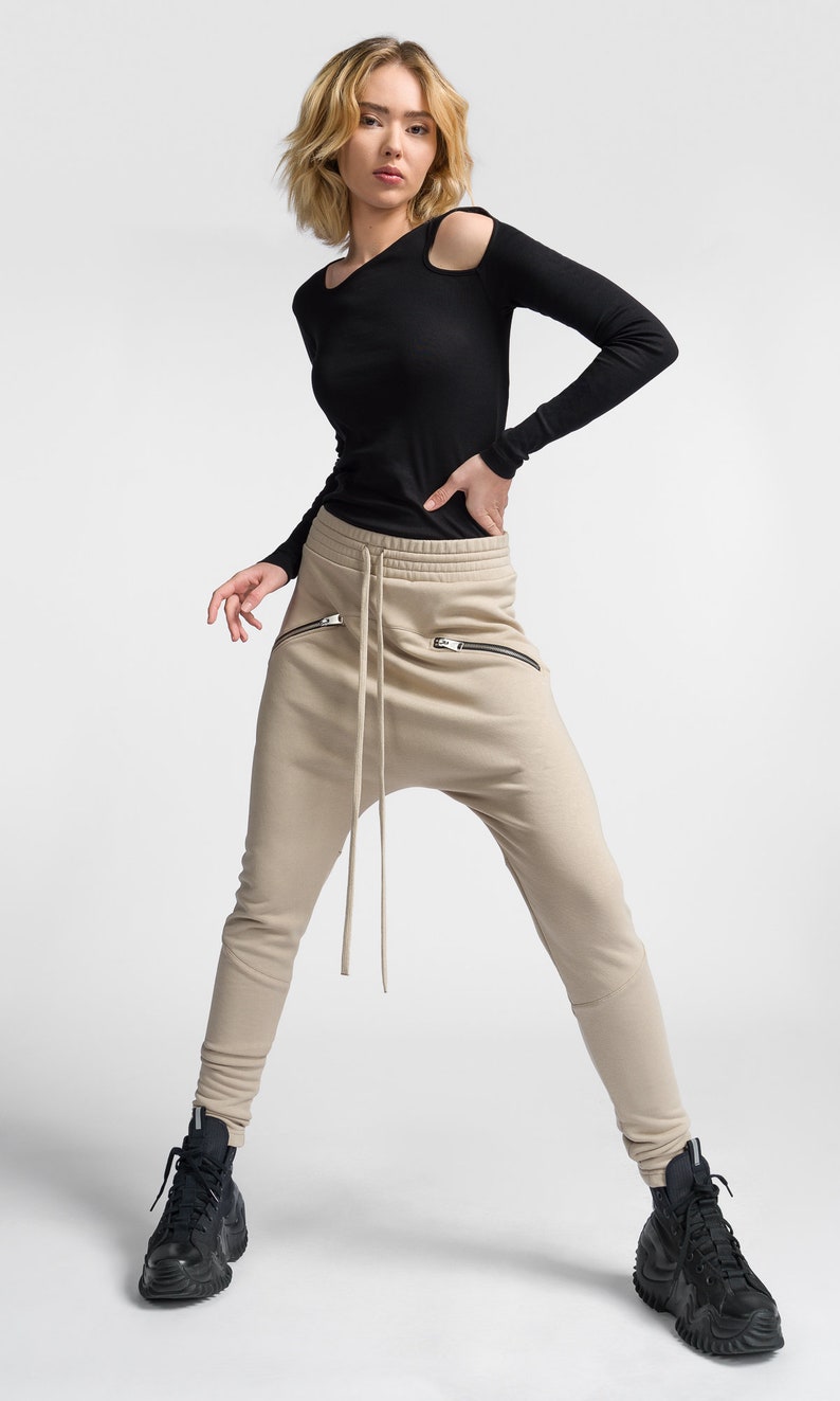 Puede incluir: Una mujer con una camisa negra de manga larga con hombros descubiertos y pantalones beige con cord&oacute;n y bolsillos con cremallera. Lleva zapatillas negras de ca&ntilde;a alta.