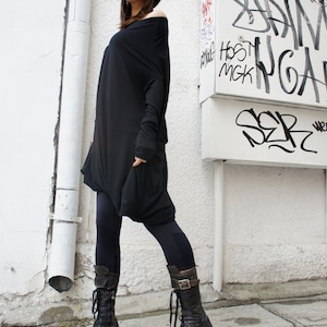 Op de afbeelding: Een vrouw draagt een zwarte off-shoulder tuniek, zwarte leggings en zwarte leren laarzen met gespen. Ze staat voor een witte muur met graffiti.