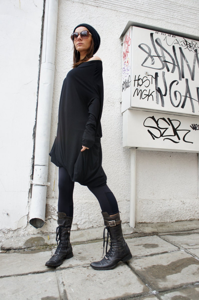 Op de afbeelding: Een vrouw draagt een zwarte off-shoulder tuniek, zwarte leggings en zwarte leren laarzen met gespen. Ze staat voor een witte muur met graffiti.