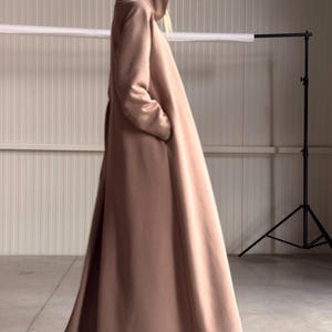 May include: A long, flowing, hooded coat in a soft, muted rose color. The coat features a full, flared skirt and is made of a smooth, possibly wool or cashmere-like fabric. The design includes pockets.