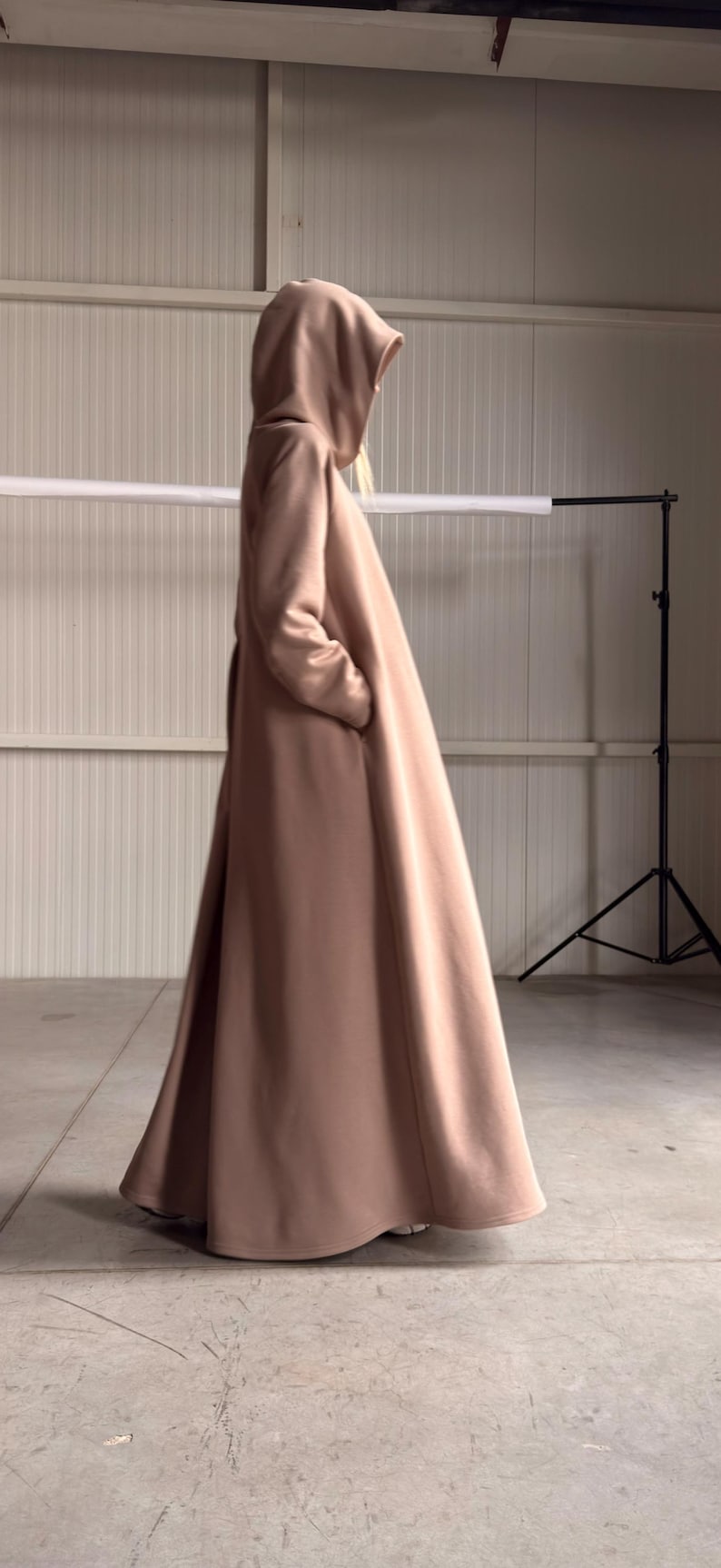 May include: A long, flowing, hooded coat in a soft, muted rose color. The coat features a full, flared skirt and is made of a smooth, possibly wool or cashmere-like fabric. The design includes pockets.