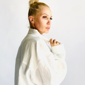 May include: A white linen shirt with long sleeves. The shirt has a classic collar and buttoned cuffs. The model is wearing the shirt and has blonde hair styled in a bun.