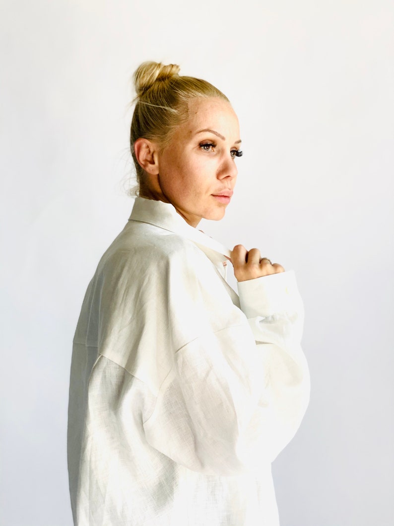 May include: A white linen shirt with long sleeves. The shirt has a classic collar and buttoned cuffs. The model is wearing the shirt and has blonde hair styled in a bun.