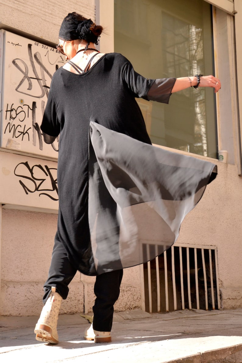 May include: A person wearing a black tunic with a sheer overlay and black pants. The tunic has a long, flowing design and the person is wearing a black headband.