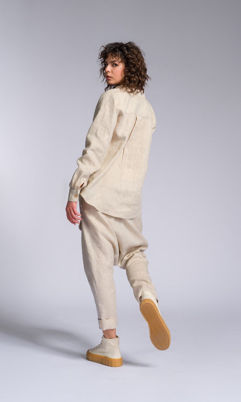 May include: A woman wearing a light beige linen shirt and pants. She is wearing white sneakers with a brown sole. The woman is standing on a white background.